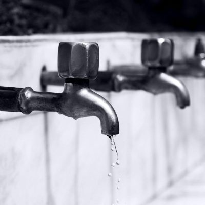 Black and white image of a row of faucets with water dripping, highlighting plumbing details.